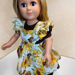 May include: A doll with blonde hair and blue eyes wearing a light blue dress with a sunflower and bird print. The dress has yellow sunflowers, brown birds, and a brown bodice. The doll is standing on a white surface.
