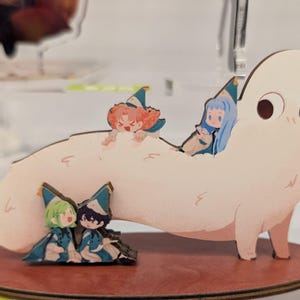 May include: A quirky wooden figurine of a large, white, cartoonish creature with big eyes and small legs. Several small, colourful anime-style characters are perched on its back. The figurine sits on a brown, circular base.