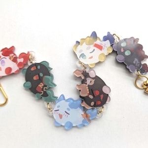 May include: Two keychains featuring colorful cartoon characters. One keychain has a red, black, and green character, and the other has a white, black, and blue character. Each keychain has a gold-colored clasp.
