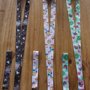 May include: Four colorful lanyards with different patterns. The lanyards are brown with a coffee cup design, white with an orange slice design, green with a kiwi design, and brown with a kiwi design. Each lanyard has a silver clip and a black string.