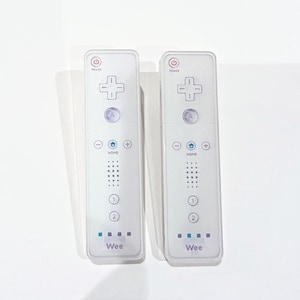 Puede incluir: Dos mandos blancos de Nintendo Wii con botones negros y un logotipo "Wii" azul y blanco. Los mandos tienen un botón de encendido, un botón de inicio y un pad direccional.