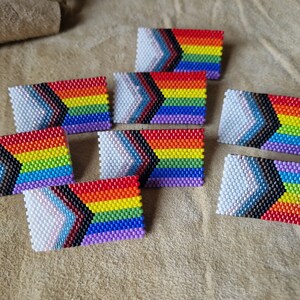 May include: Eight beaded rainbow pride flags with a black and brown chevron design. The flags are white, purple, blue, green, yellow, orange, and red.