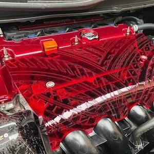 Puede incluir: Tapa de motor roja con un diseño estampado. La tapa está hecha de plástico transparente con un tinte rojo. La tapa está asegurada con tornillos plateados.