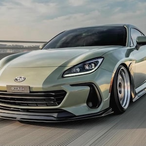 Op de afbeelding: Een saliegroene sportwagen met een strak design wordt in beweging vastgelegd op een snelweg. De auto heeft een laag profiel, zwarte accenten en de tekst "BRZCRUSH" op de voorkant. De wielen hebben een zilveren afwerking.