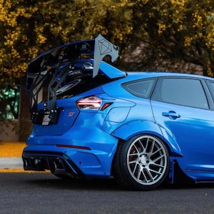 Könnte beinhalten: Ein blauer Ford Focus ST mit einem großen schwarzen Heckspoiler und Aftermarket-Felgen. Das Auto hat ein tiefergelegtes Fahrwerk und eine kundenspezifische Auspuffanlage.