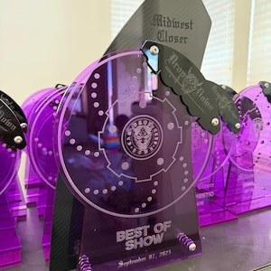Op de afbeelding: Een verzameling paarse acryl prijzen. Elke prijs heeft een cirkelvormig ontwerp met een tandwielachtig patroon, een zwart accentstuk en de tekst "Midwest Closer" en "Best of Show September 07, 2023".