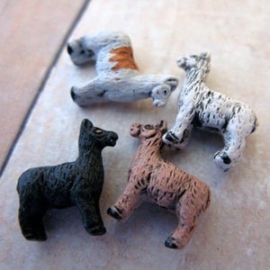May include: Four small ceramic llama figurines. One is black, one is white, one is brown and white, and one is brown. The figurines are all standing on a light brown surface.