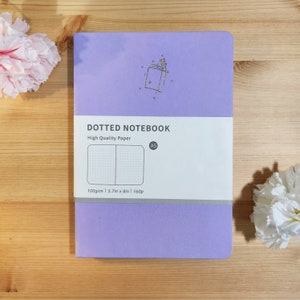 May include: A lavender dotted notebook with a gold book and stars design. The notebook is A5 size and has 100gsm paper. The text on the front reads "DOTTED NOTEBOOK High Quality Paper A5 100gsm | 5.7in x 8in | 160p"