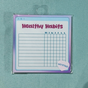 May include: A white and blue notepad with a pink heart sticker that says "NOTEPAD". The notepad has a grid pattern and the words "Healthy Habits" at the top. The notepad has a rainbow gradient background.