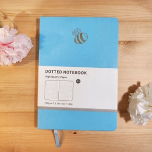 May include: A light blue A5 dotted notebook with a gold bee design. The cover reads "DOTTED NOTEBOOK" and "High Quality Paper". The notebook is 100gsm and measures 5.7in x 8in. It has 160 pages.