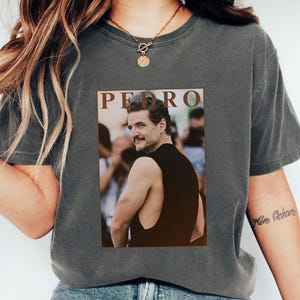 Peut inclure: T-shirt gris foncé avec une image rectangulaire. L'image présente le nom "PEDRO" au-dessus d'une photo d'un homme en débardeur noir. Le t-shirt est de style décontracté.