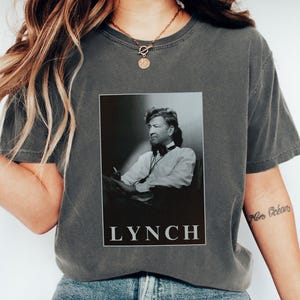 May include: A charcoal grey t-shirt with a black and white image of David Lynch, a filmmaker, wearing a white shirt and tie. The text "LYNCH" is printed in white below the image.