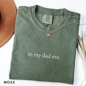 Peut inclure: T-shirt vert mousse avec l'inscription blanche "in my dad era". Un collier doré avec un pendentif repose sur le t-shirt. Le t-shirt est plié et posé sur une surface blanche. Un chapeau marron est visible en arrière-plan.