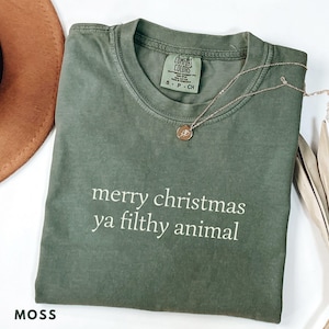 May include: A moss green t-shirt with the phrase "merry christmas ya filthy animal" printed in white. The shirt has a crew neck and short sleeves. A gold necklace with a small pendant is draped across the shirt.