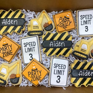 May include: A box of decorated sugar cookies with a construction theme. Cookies include road signs that say "Party Zone" and "Speed Limit 3", construction vehicles, and cookies with the name "Aiden". The cookies are yellow, black, and white.