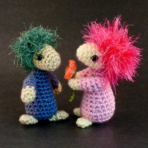 May include: Two crocheted amigurumi figures, one with green hair and a blue outfit, the other with pink hair and a pink outfit, holding a red flower.