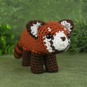 May include: A crocheted red panda plush toy with brown legs and a white face. The toy has black eyes and a small pink nose.