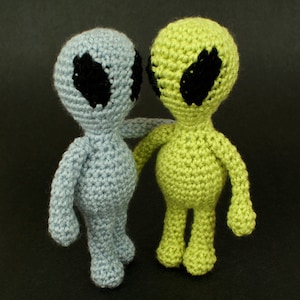 Puede incluir: Dos juguetes extraterrestres de crochet, uno azul y otro verde, con ojos negros. Los extraterrestres están de pie uno al lado del otro y parecen mirarse.