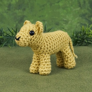 Peut inclure: Un jouet en crochet représentant un lionceau de couleur brun clair avec des yeux noirs. Le jouet est debout sur une surface verte.