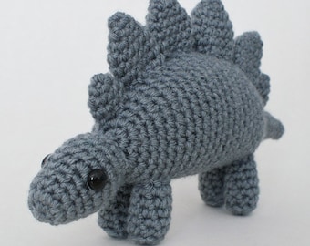May include: A gray crocheted dinosaur toy with black eyes. The dinosaur has a spiky back and a long tail.