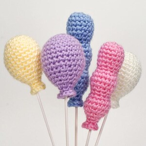 May include: Five crocheted balloons in various pastel colors, including yellow, purple, blue, pink, and white, are attached to white sticks. The balloons are all in a bunch, and the sticks are all pointing upwards.
