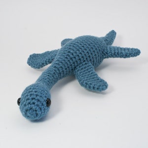 Op de afbeelding: Een gehaakt blauw pluche speelgoed in de vorm van een plesiosaurus, een langhalzige zeereptiel. Het speelgoed heeft zwarte ogen en een lange nek.