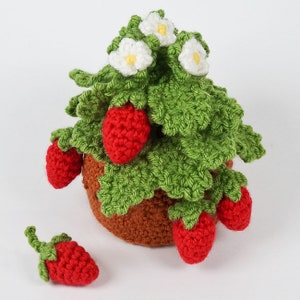 Puede incluir: Una planta de fresas de crochet en una maceta marrón con fresas rojas y flores blancas. La planta está sobre una superficie blanca.