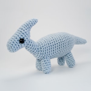 May include: A light blue crocheted dinosaur toy with black eyes. The dinosaur is standing on a white surface.