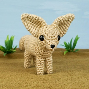May include: A crocheted fennec fox toy with large ears, black eyes, and a brown nose. The toy is standing on a brown surface.