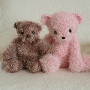 May include: Two plush teddy bears, one brown and one pink, are sitting on a white surface. Both bears have black eyes and a black nose.