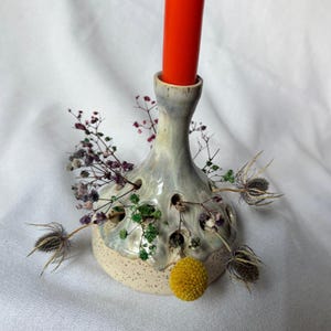 Peut inclure: Un bougeoir en céramique avec une base tachetée et un corps bleu-gris clair. Le bougeoir comporte plusieurs trous et est décoré de fleurs séchées et d'un pompon jaune. Une bougie orange est insérée en haut.