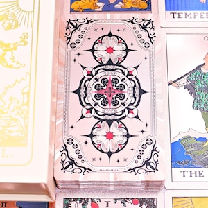 May include: A deck of tarot cards featuring a cream-colored box with a gold foil illustration of The Fool. The card backs display a symmetrical black, white, and pink floral pattern. Several individual tarot cards are visible, including The Fool.