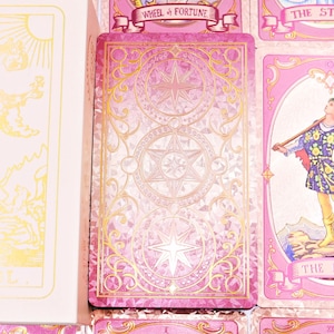 May include: A pink tarot card deck with gold accents. The cards feature illustrations and text, including "The Fool" and "Wheel of Fortune." The deck is displayed with a matching box, showcasing a golden illustration of a figure.