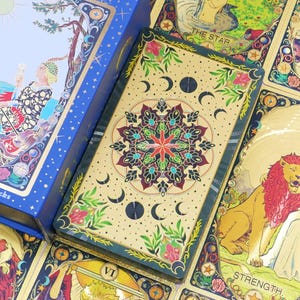 Puede incluir: Una caja azul y una baraja de cartas del tarot con ilustraciones y texto. La caja dice "a. weite Tarot deck" y "78 latest Waite Taro decks." Las cartas tienen bordes dorados y representan varias figuras y símbolos.