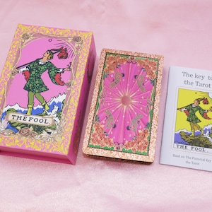 May include: A pink tarot card box and deck with a matching booklet. The box features an illustration of a figure in green clothing, labeled "THE FOOL." The deck has a pink background with a central sun design. The booklet is titled "The key to the Tarot."