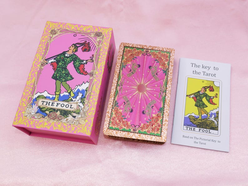 May include: A pink tarot card box and deck with a matching booklet. The box features an illustration of a figure in green clothing, labeled "THE FOOL." The deck has a pink background with a central sun design. The booklet is titled "The key to the Tarot."