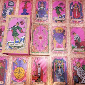 May include: A pink tarot card deck with a matching box, featuring illustrations of various figures and symbols. The cards are arranged on a pink surface, showcasing the deck's design. The box displays "THE FOOL" on the front.