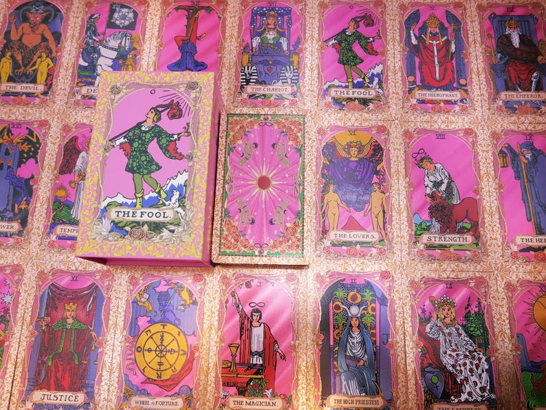 May include: A pink tarot card deck with a matching box, featuring illustrations of various figures and symbols. The cards are arranged on a pink surface, showcasing the deck's design. The box displays "THE FOOL" on the front.