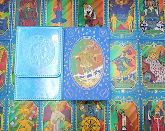 Puede incluir: Una caja de cartas de tarot holográfica azul y una caja rectangular azul a juego con una ilustración ovalada central de una figura con túnica estampada, rodeada de símbolos del zodiaco y estrellas doradas sobre un fondo de cartas de tarot.