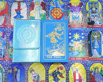 May include: A light blue iridescent deck box with a circular embossed design next to a blue tarot card box featuring a gold foil illustration of The Fool and the text THE FOOL, set against a background of various tarot card illustrations.