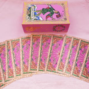 May include: A pink tarot card deck with a matching box. The box features an illustration of a figure labeled "THE FOOL." The cards are fanned out, displaying pink and gold designs with floral accents. The overall aesthetic is whimsical and colorful.