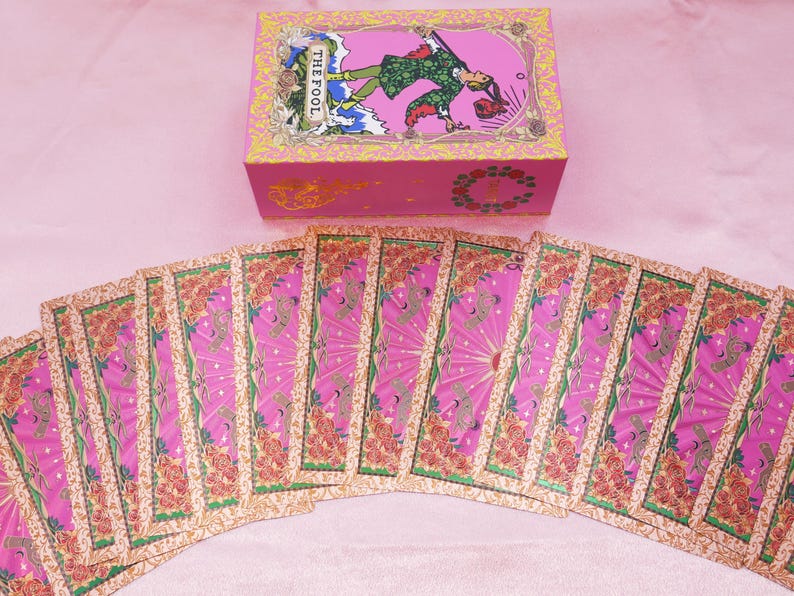 May include: A pink tarot card deck with a matching box. The box features an illustration of a figure labeled "THE FOOL." The cards are fanned out, displaying pink and gold designs with floral accents. The overall aesthetic is whimsical and colorful.