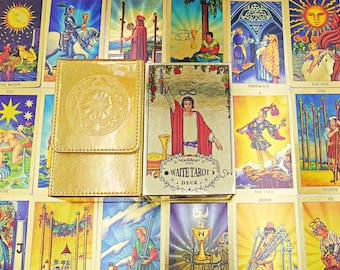 Puede incluir: Una caja de cartas de tarot de color dorado con cierre de solapa magnética se encuentra junto a una caja de cartas de tarot dorada a juego que muestra una figura con túnica roja sosteniendo una varita, con el texto Golden WAITE TAROT DECK.