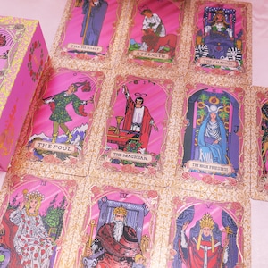 May include: A pink tarot card deck with gold floral accents and individual cards laid out. The cards feature illustrations of The Fool, The Magician, The High Priestess, and other major arcana. The box is also pink with gold floral details.