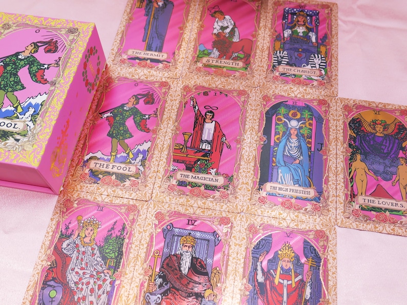 May include: A pink tarot card deck with gold floral accents and individual cards laid out. The cards feature illustrations of The Fool, The Magician, The High Priestess, and other major arcana. The box is also pink with gold floral details.