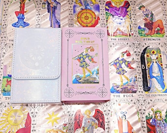 Puede incluir: Una caja de almacenamiento para cartas de tarot de color azul claro iridiscente junto a una caja de mazo de tarot rosa con el título Gold Foil Tarot y una ilustración del personaje El Loco con detalles en lámina dorada.