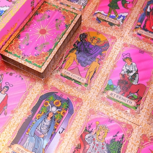 May include: A pink tarot card deck with gold accents and a matching box. The cards feature illustrations with titles like "The Lovers" and "Strength." The deck is set against a floral patterned background.