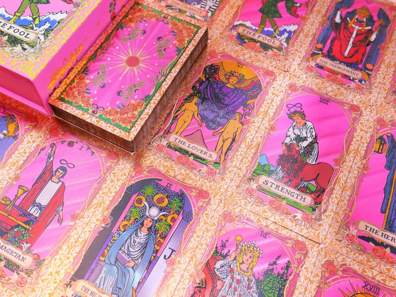 May include: A pink tarot card deck with gold accents and a matching box. The cards feature illustrations with titles like "The Lovers" and "Strength." The deck is set against a floral patterned background.