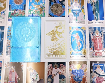 May include: A metallic light blue deck box with a circular embossed design next to a white tarot card box featuring a gold foil illustration of The Fool. The items are placed on a background of various tarot cards with blue and gold accents.