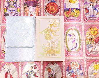 Può includere: Un cofanetto per tarocchi rosa chiaro con un'illustrazione in lamina d'oro della carta Il Matto e una custodia argentata dalla texture metallica con un motivo circolare in rilievo, entrambi su uno sfondo di varie carte.